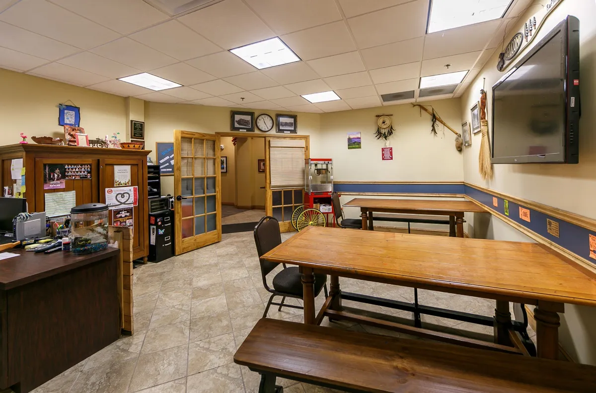 A welcoming interior scene with wooden tables set for dining, a front desk visible in the background, and tasteful wall decorations. The mood feels friendly and communal, ideal for gatherings and shared meals.