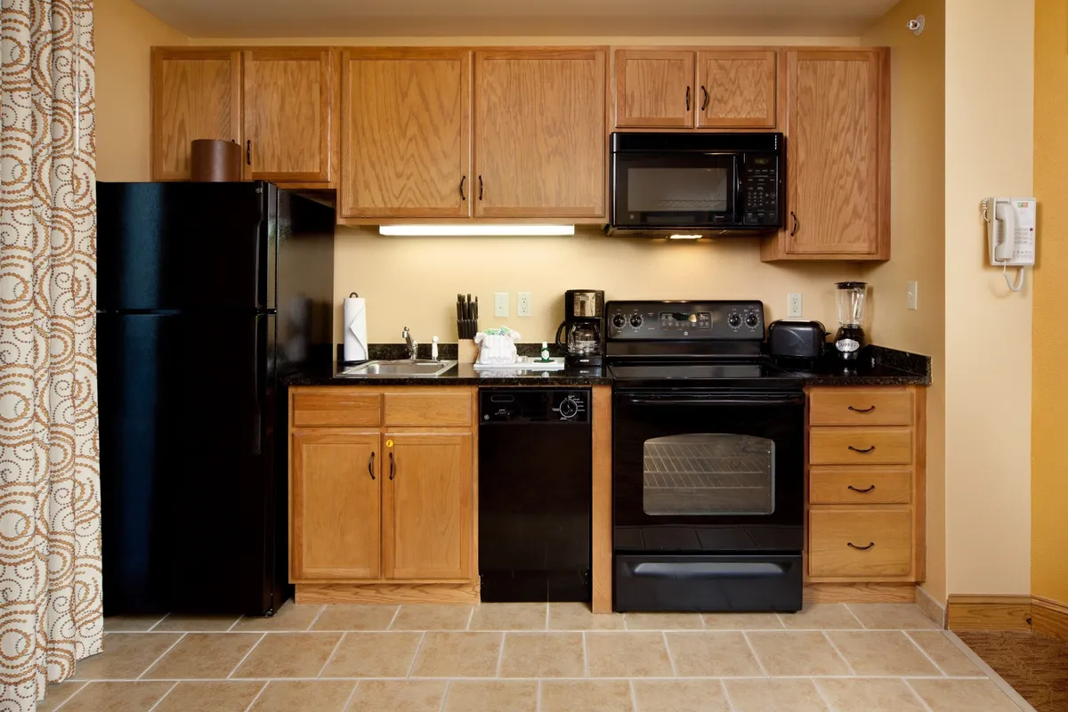 This image captures the condo kitchen, featuring sleek black appliances, warm wood cabinets, a granite countertop, and a stainless steel sink. The setting is cozy and functional, with signs of meal preparation and family activity, such as children baking cookies and adults cooking meals together.