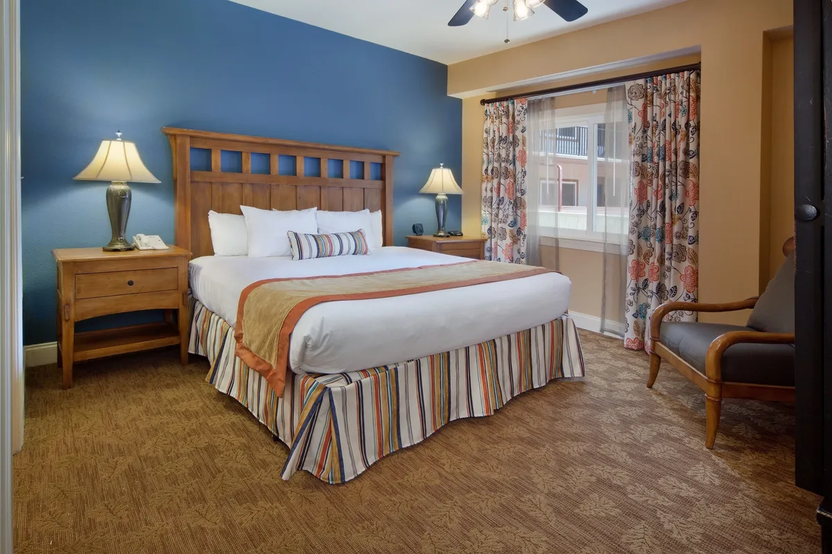 This image features a king bed with decorative pillows, wooden nightstands with lamps, cozy curtains, and an accent chair by the window. The natural light flows into the room, creating a restful and inviting environment perfect for unwinding.