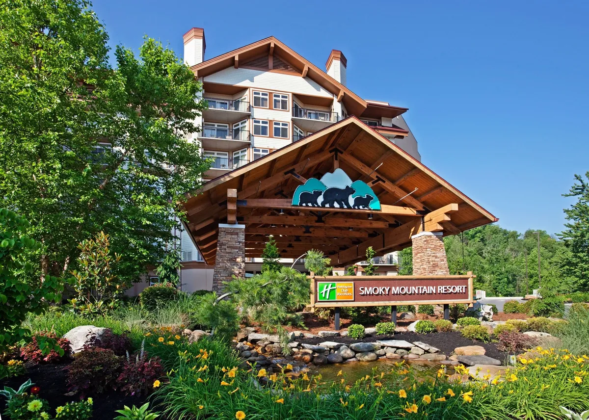 This exterior shot captures the welcoming entrance canopy and hotel sign, set amidst beautifully landscaped gardens with sweeping views of the surrounding mountains. Families and guests can be seen arriving and relaxing outdoors, hinting at the lodge's inviting atmosphere and the promise of shared experiences in nature.