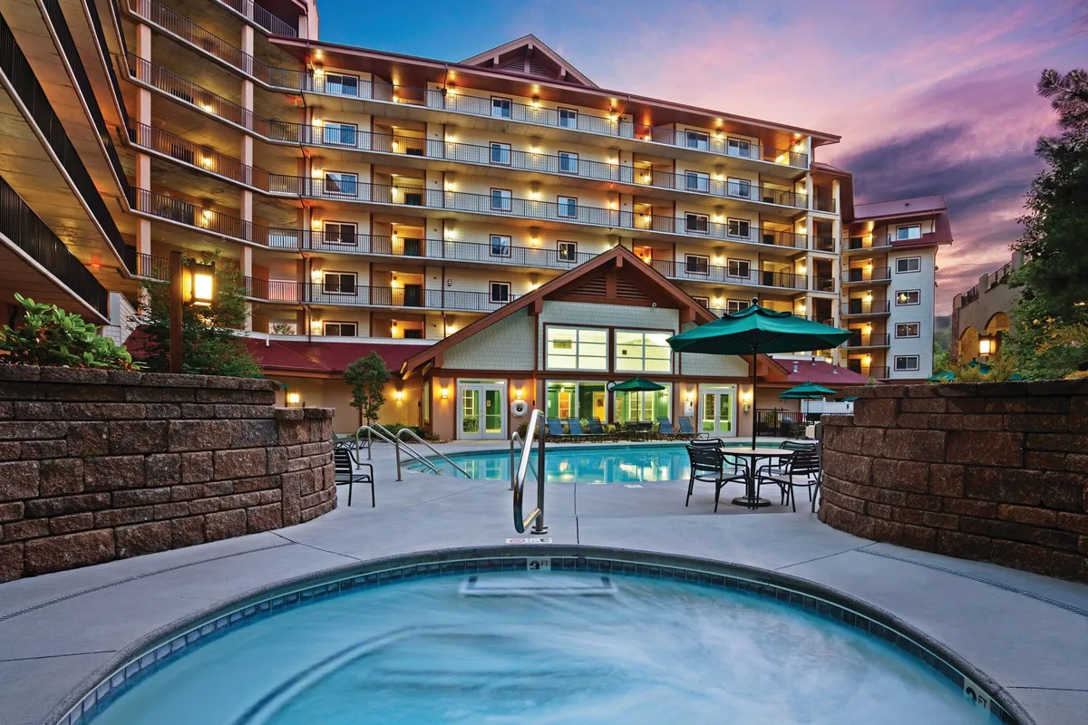 This exterior image showcases a lively outdoor space at dusk, featuring a pool, a hot tub, and nearby seating areas. A group enjoys relaxed evening activities around a fire pit, with the lobby entrance softly illuminated in the background. The setting captures guests gathering, socializing, and unwinding as the sun sets behind the Smoky Mountains, blending the natural scenery with the warmth of communal amenities.