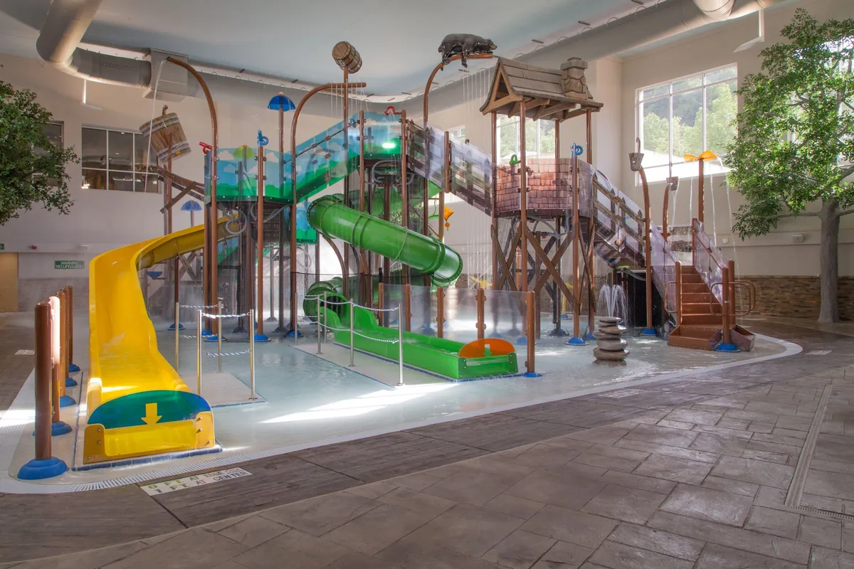 A vibrant indoor water park filled with colorful water slides, a playful splash structure, and whimsical water features. Children are laughing and playing, while families gather around watching and participating. The space is bright, lively, and visually exciting, filled with movement and energy.