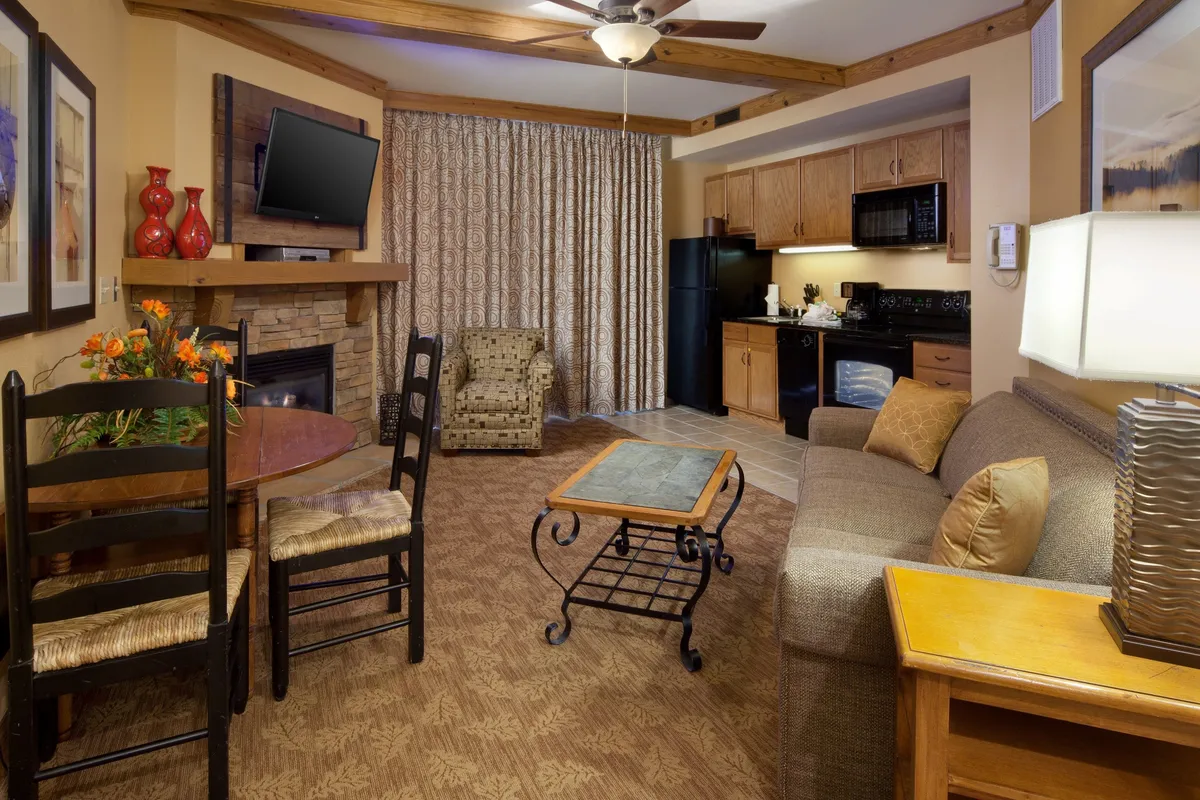 This inviting hotel condo living space features a cozy fireplace, a plush sofa for relaxing, a family-sized dining table, a large television for movie nights, and an open kitchen area—all bathed in warm, welcoming light.