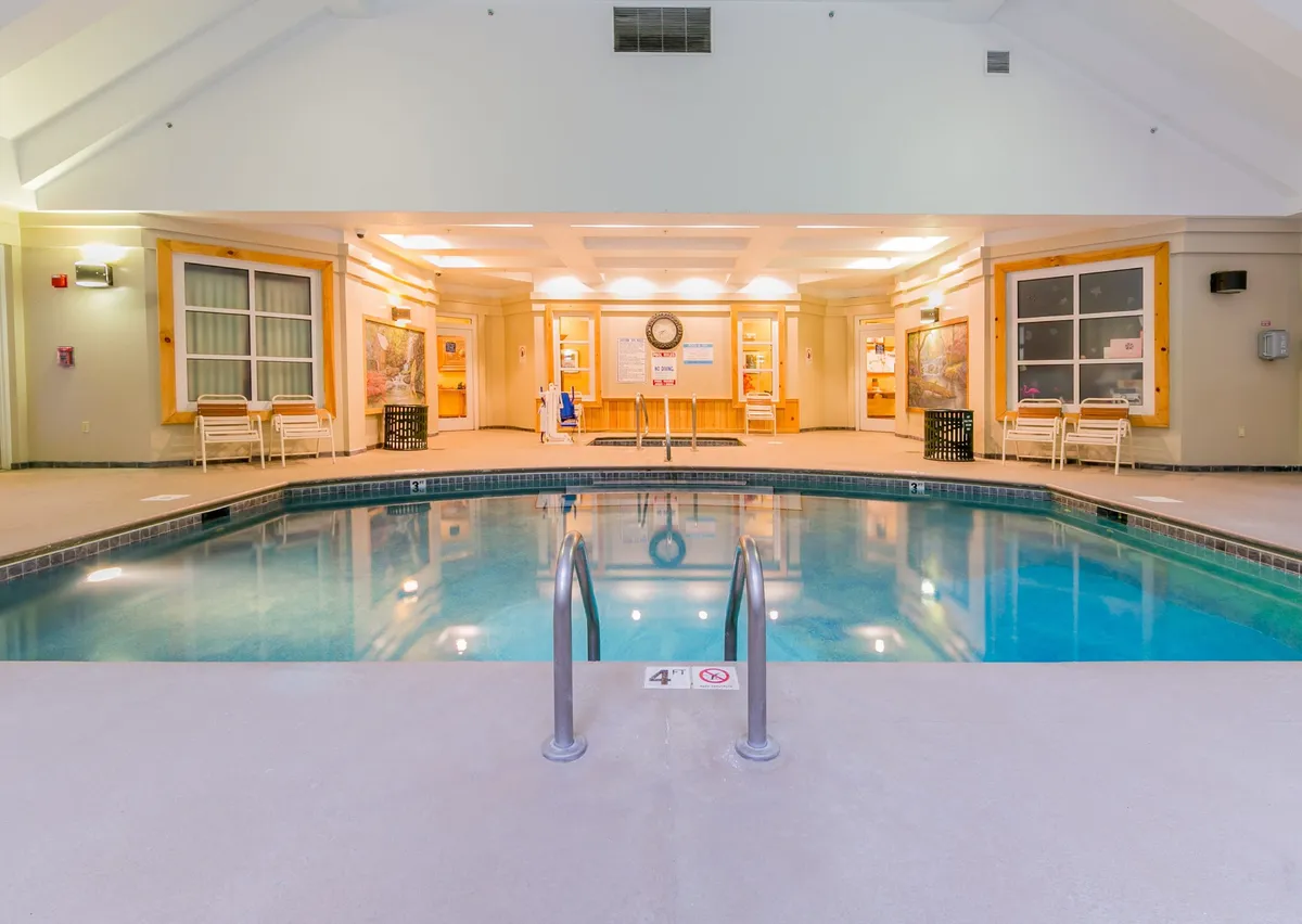 An inviting indoor swimming pool area featuring wide pool steps, comfortable lounge chairs lining the edges, and stylish decorative wall accents. The setting is calm yet ideal for swimming, relaxing, and socializing, with natural light creating a welcoming atmosphere.